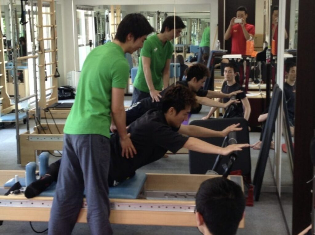 ピラティススタジオBB 錦糸町のクリニカルリフォーマーで、国松亜樹良インストラクターがAce pilates Studioの金城インストラクターにマーメイドツイストのエクササイズを指導している写真