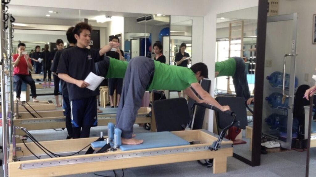 ピラティススタジオBB 錦糸町のクリニカルリフォーマーで、Ace pilates Studioの金城インストラクターが国松亜樹良インストラクターにエレファントのエクササイズを指導している写真