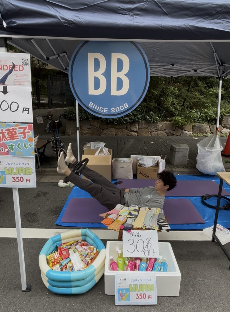 恵比寿「ビール坂祭り」にて、ピラティススタジオBB恵比寿店の出店様子。
ピラティススタジオBB男性スタッフが、「ハンドレッド」というエクササイズを行なっている。お祭りで販売したピラティス専用靴下や、MURB(プロテインドリンク)、駄菓子掬いの様子もわかる。