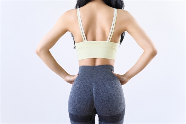 白い背景の前で、黄緑色のブラトップと濃いグレーのレギンスを着用した女性の引き締まった背中の後ろ姿