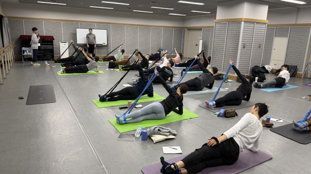 ViktorUyganが東京のコネクションズバンドを用いたセミナーでマットエクササイズのサイドキックを指導しているシーン