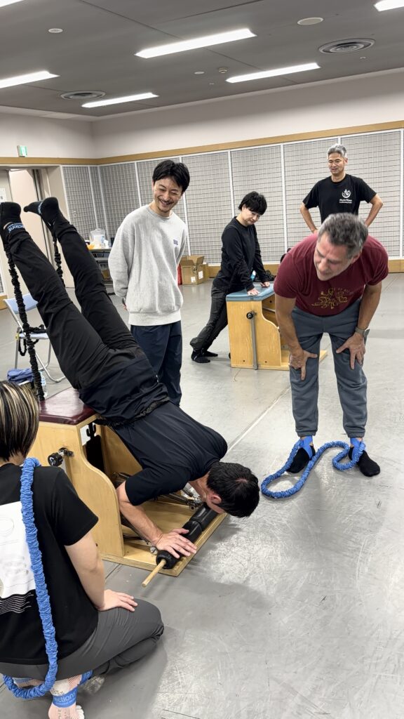 ViktorUyganと通訳、ピラティススタジオBBのインストラクターが、チェアでコネクションズバンドを用いたスワンダイブを実施している画像