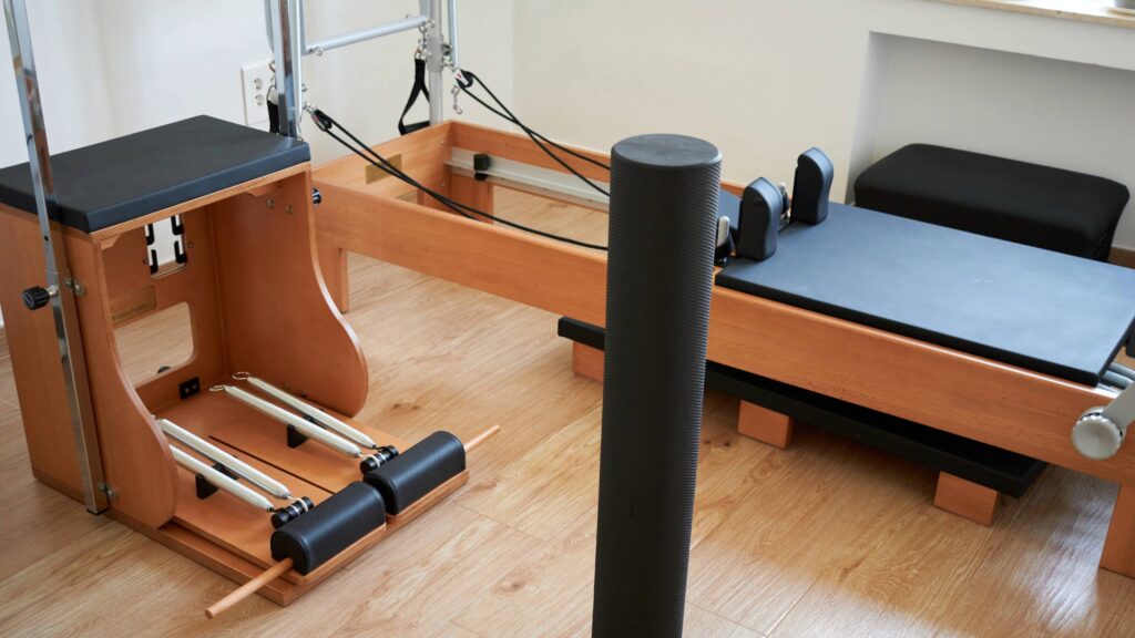 木目調の床に、スプリングの付いたピラティスチェアとリフォーマー、手前に黒いフォームローラーが置かれた室内。