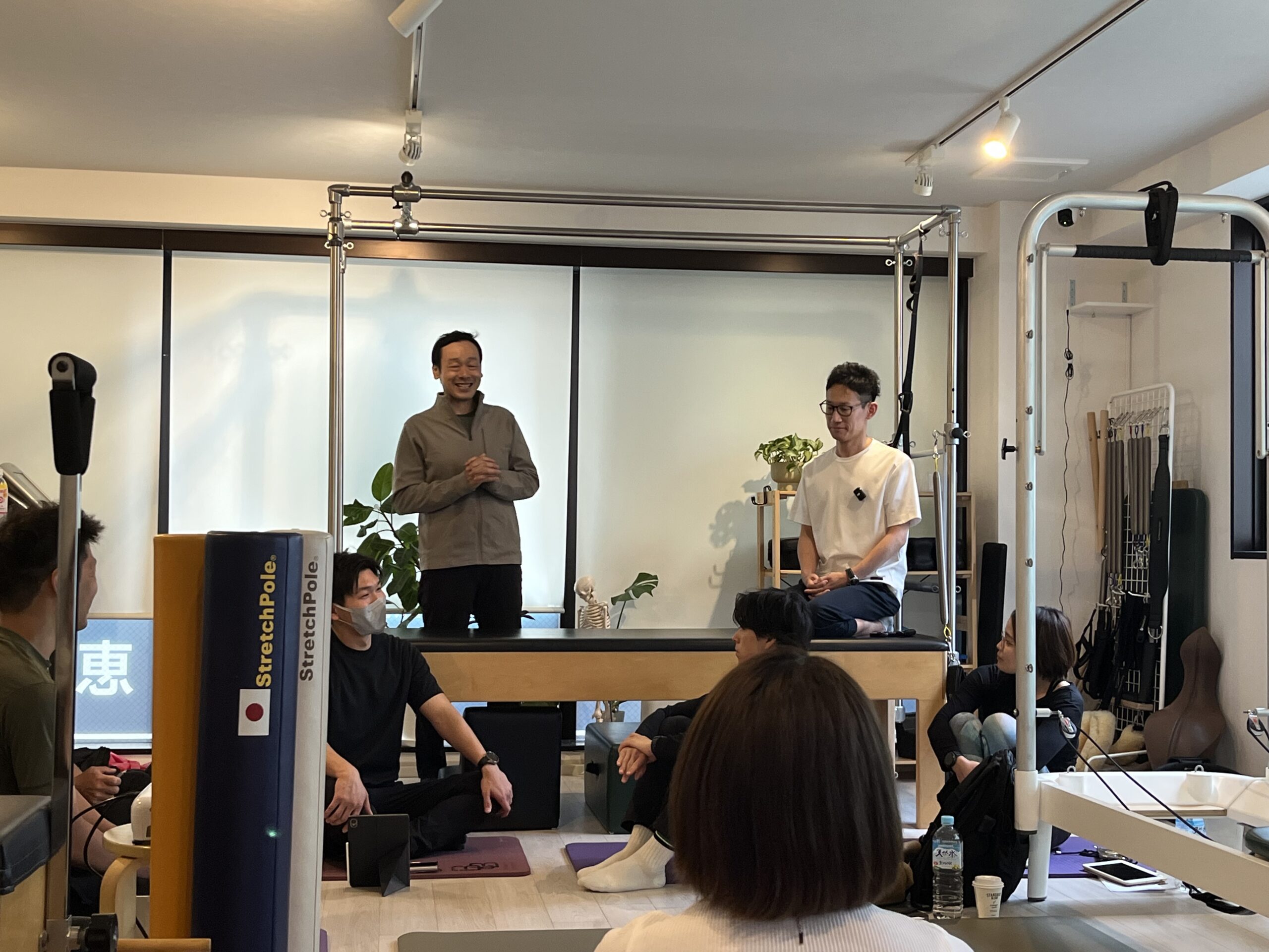 Two instructors lead a small group in a gym or studio; one man stands and speaks while another sits, with attendees seated on mats nearby.